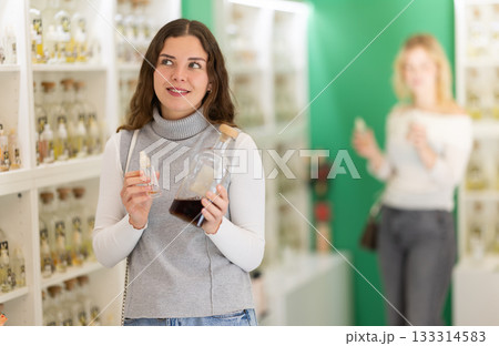 Girl sniffs fragrance of perfume, chooses scent spray, woman shopping in background 133314583