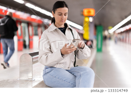 Girl with phone is sitting on metro platform station, scrolling though internet Girl with phone is sitting on metro platform station, scrolling though internet 133314749