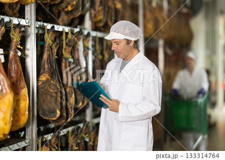 In jamon production shop, man employee inspects finished product and fills out documents In jamon production shop, man employee inspects finished product and fills out documents 133314764
