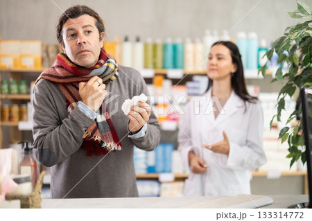 Man in scarf with sore throat is looking for medicine in pharmacy. Female pharmacist is helping him Man in scarf with sore throat is looking for medicine in pharmacy. Female pharmacist is helping him 133314772