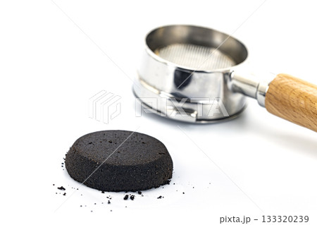 Macro shot of a compressed coffee puck next to a stainless steel portafilter with a wooden handle on a white background Macro shot of a compressed coffee puck next to a stainless steel portafilter with a wooden handle on a white background 133320239