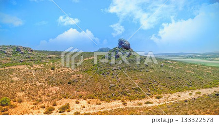 Rocky hill topped by boulder is rising in sunlit semi-arid landscape, with sparse shrubs 133322578