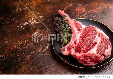 Fresh Tomahawk beef steak, raw marbled rib eye with bone on plate with herbs. brown background. top view Fresh Tomahawk beef steak, raw marbled rib eye with bone on plate with herbs. brown background. top view 133323655