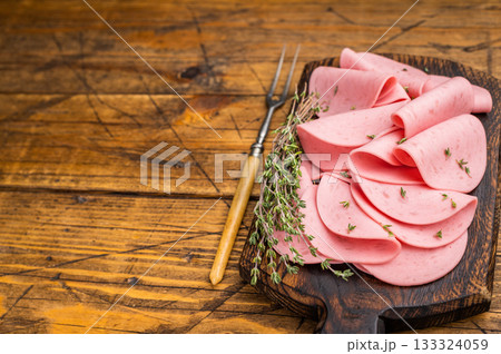 Slices of pink bologna lunch meat served on a rustic dark wooden cutting board. Dark, moody background with copy space. Slices of pink bologna lunch meat served on a rustic dark wooden cutting board. Dark, moody background with copy space. 133324059