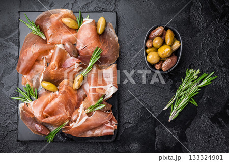 Jamon Iberico (Spanish cured ham) and olives on a black slate board. Top view with large copy space on a dark textured background. 133324901