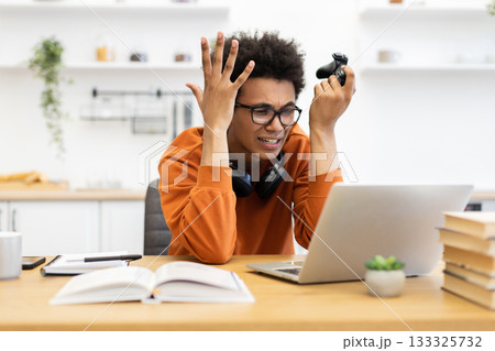 A young man is visibly frustrated while playing a video game on his laptop, holding a game controller. 133325732