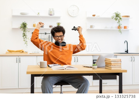 A young man with glasses is enthusiastically celebrating a win while gaming at his desk in a bright kitchen setting. 133325738