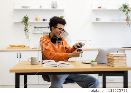 A young man with glasses is smiling while playing video games on his laptop at a wooden table. 133325751