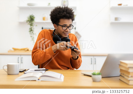 A young man with glasses is engrossed in a video game, holding a controller while sitting at a desk with a laptop and books. 133325752