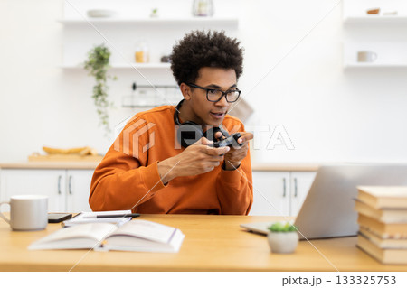 A young man with glasses is engrossed in a video game, holding a controller while sitting at a table with a laptop. A young man with glasses is engrossed in a video game, holding a controller while sitting at a table with a laptop. 133325753
