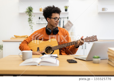A young man with glasses plays an acoustic guitar while looking at a laptop in a bright kitchen setting. A young man with glasses plays an acoustic guitar while looking at a laptop in a bright kitchen setting. 133325770