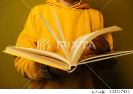 Person in yellow hoodie reading open book against warm brown background. a woman reading at home. A person's hand is holding an open book with slightly yellowed paper pages Person in yellow hoodie reading open book against warm brown background. a woman reading at home. A person's hand is holding an open book with slightly yellowed paper pages 133327480