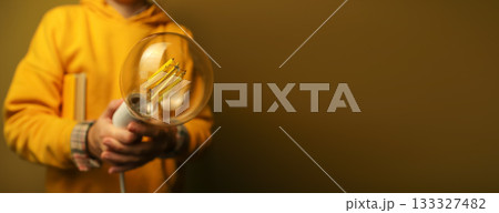 Person in yellow hoodie holding large glowing vintage-style LED light bulb against warm background. Hand holding a filament light bulb isolation on dark yellow background. Innovation concept 133327482