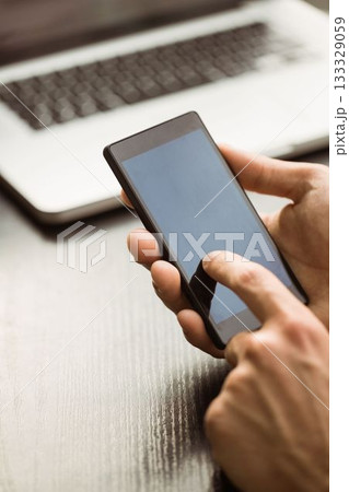 Hands are holding black smartphone and tapping screen above laptop on wooden desk in flat design Hands are holding black smartphone and tapping screen above laptop on wooden desk in flat design 133329059