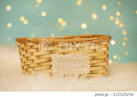 A natural wicker basket with a lace-edged Home label stands on a fluffy white surface against a soft blue background with warm bokeh lights. comfort, coziness, and home decor aesthetics 133329250