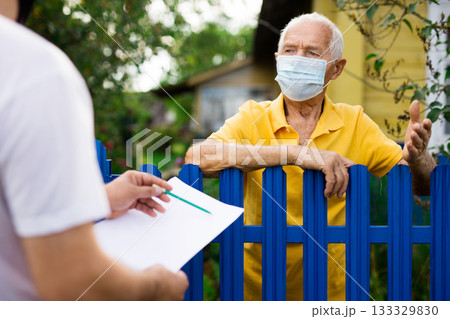 Grandfather in protective mask talking to census agent standing at fence of his country house 133329830