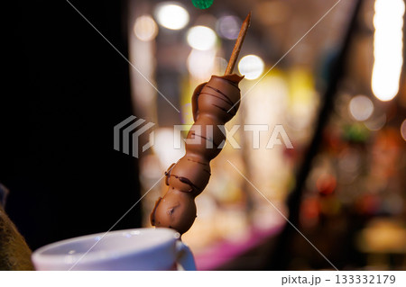 Chocolate dessert at a night Christmas market. Choco covered grape skewer with soft bokeh lights in the background,  creating a warm, festive atmosphere, 133332179