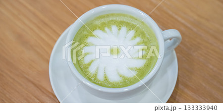 A warm cup of matcha latte rests on a white saucer atop a wooden table, with white foam forming a simple floral design in the center. A warm cup of matcha latte rests on a white saucer atop a wooden table, with white foam forming a simple floral design in the center. 133333940