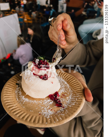 Woman holding disposable plate and eating meringue dessert with whipped cream, pomegranate seeds, mint and powdered sugar, inside crowded place. Woman enjoying meringue dessert with whipped cream Woman holding disposable plate and eating meringue dessert with whipped cream, pomegranate seeds, mint and powdered sugar, inside crowded place. Woman enjoying meringue dessert with whipped cream 133336199