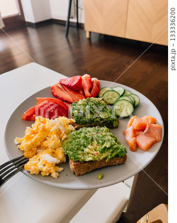 Grey plate with wholemeal avocado toast, scrambled eggs, smoked salmon, sliced tomatoes, strawberries and cucumbers served for breakfast on white table. Wholemeal avocado toast with scrambled eggs 133336200