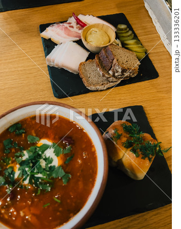 Ukrainian soup with sour cream and herbs, accompanied by bread and platter of salo, rye bread, pickled cucumbers, mustard, and chili pepper, served on black slate plates. Traditional Ukrainian meal 133336201
