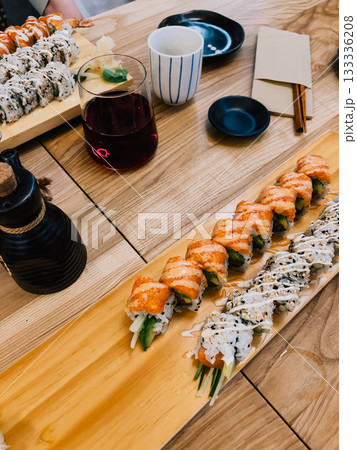 Wooden table hosting delightful assortment of sushi rolls, accompanied by wine, soy sauce, ginger, wasabi, and cup of tea, creating vibrant and inviting dining experience. Colorful sushi rolls 133336208