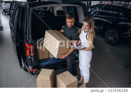 Woman is helping delivery man with products, holding notepad, loader is working with boxes from automobile 133336288