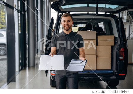 Pen and the notepad in hands. Delivery man or loader is near the car with a box Pen and the notepad in hands. Delivery man or loader is near the car with a box 133336465