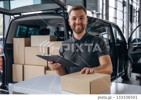 Positive facial expression, holding notepad. Delivery man or loader is near the car with a box 133336501