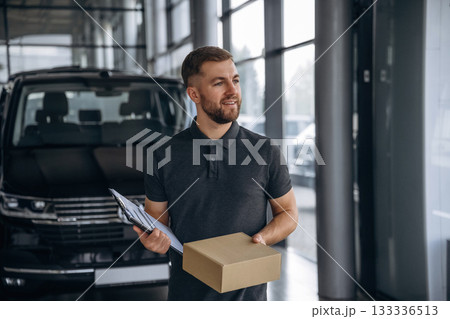 Standing, holding notepad. Delivery man or loader is near the car with a box Standing, holding notepad. Delivery man or loader is near the car with a box 133336513
