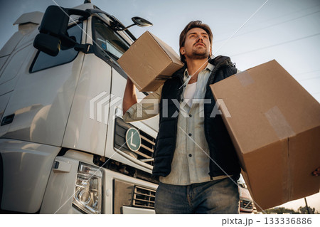 Looking forward, holding two boxes. Truck driver is near the transport outdoors Looking forward, holding two boxes. Truck driver is near the transport outdoors 133336886