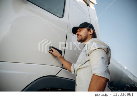 In the casual clothes and in black cap. Truck driver is near the transport outdoors In the casual clothes and in black cap. Truck driver is near the transport outdoors 133336961
