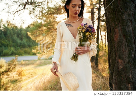 White dress. Bouquet in hands. Woman is outdoors in nature with bouquet of flowers White dress. Bouquet in hands. Woman is outdoors in nature with bouquet of flowers 133337384