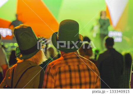 Couple in green hats enjoying St. Patrick Day celebration at club with Irish flag lights on stage. Concept of unity, tradition, national pride 133340915