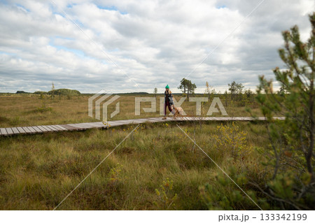 Thoughtful woman and hunting dog walking along boardwalk on peat bog, trail under cloudy sky 133342199