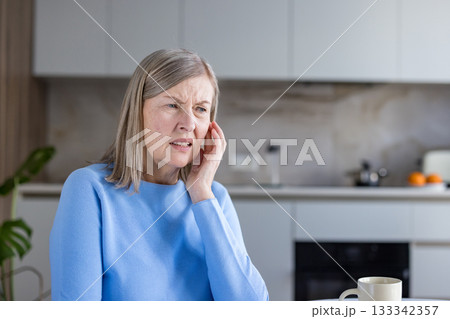 Senior woman suffering from a painful earache, touching her ear with a distressed expression while experiencing an uncomfortable health problem at home in her kitchen 133342357