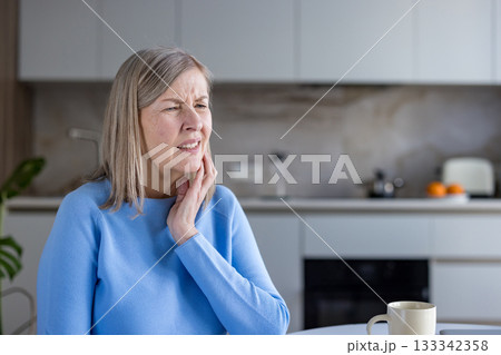 Mature woman feeling intense pain in her jaw or neck, touching her cheek and clenching her teeth, indicating discomfort and need for relief or medical attention 133342358