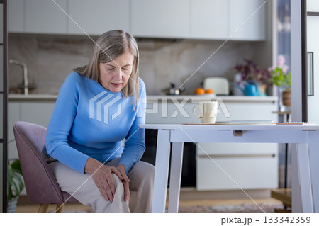 Senior woman sitting in her kitchen gripping her painful knee, showing discomfort from age-related joint pain and arthritis affecting everyday home life and mobility 133342359