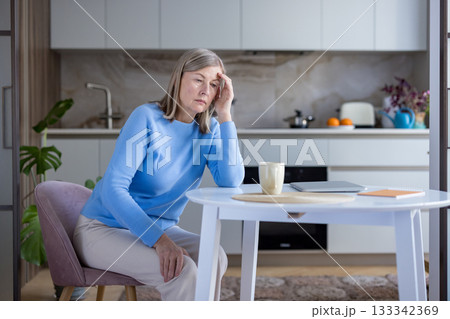 Mature woman sitting at a kitchen table experiencing a stressful headache, feeling tired and lonely while thinking about her depression and seeking comfort at home Mature woman sitting at a kitchen table experiencing a stressful headache, feeling tired and lonely while thinking about her depression and seeking comfort at home 133342369