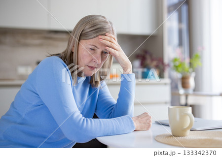 Senior grey-haired woman seated at a kitchen table looking down with hand on forehead, appearing pensive and distressed, suffering from headache, worry and loneliness Senior grey-haired woman seated at a kitchen table looking down with hand on forehead, appearing pensive and distressed, suffering from headache, worry and loneliness 133342370