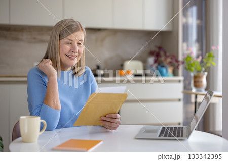 Senior woman smiling happily while reading an exciting document or letter, sitting at a table with a laptop, reflecting joy and positive emotions at home Senior woman smiling happily while reading an exciting document or letter, sitting at a table with a laptop, reflecting joy and positive emotions at home 133342395