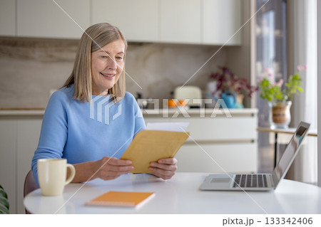 Senior woman smiling while unpacking and reading a letter or important document at a laptop-equipped table, managing paperwork at her home office kitchen Senior woman smiling while unpacking and reading a letter or important document at a laptop-equipped table, managing paperwork at her home office kitchen 133342406