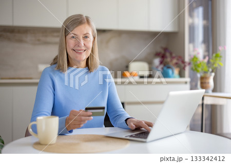 Smiling senior woman in a cozy kitchen using a laptop and holding a credit card for online shopping and banking, relaxed at the counter with a mug and natural light Smiling senior woman in a cozy kitchen using a laptop and holding a credit card for online shopping and banking, relaxed at the counter with a mug and natural light 133342412