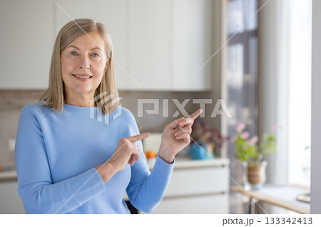 Senior woman smiling and pointing to the side in a modern kitchen, presenting empty copy space for advertisement or information, cheerful and relaxed domestic portrait Senior woman smiling and pointing to the side in a modern kitchen, presenting empty copy space for advertisement or information, cheerful and relaxed domestic portrait 133342413