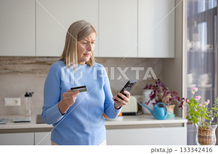 Senior woman in a modern kitchen looks confused and worried holding a credit card and smartphone while attempting an online payment, illustrating digital literacy challenges Senior woman in a modern kitchen looks confused and worried holding a credit card and smartphone while attempting an online payment, illustrating digital literacy challenges 133342416