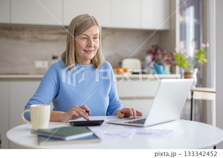 Mature woman at kitchen table managing household finances balancing bills, calculator and laptop, focused on budgeting, retirement planning and online accounting in a comfortable home setting 133342452