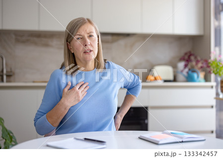 Senior woman at home experiencing a sudden chest pain and discomfort, holding her hand to her chest, possibly indicating a symptom of heart attack or heart disease 133342457