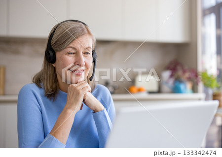 Happy senior woman with gray hair wearing a headset, smiling and working on a laptop in her bright home kitchen during a video call or remote work session 133342474