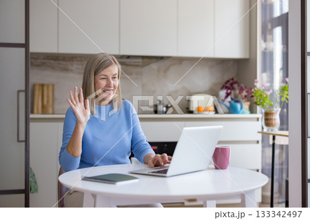 Senior woman actively engaging in a video call with a laptop from her home kitchen, enjoying modern communication technology to stay connected with family and friends Senior woman actively engaging in a video call with a laptop from her home kitchen, enjoying modern communication technology to stay connected with family and friends 133342497