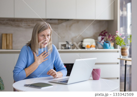 Senior woman suffering from sudden pain or anxiety, touching her chest and looking worriedly at the laptop screen during a virtual conversation in her kitchen Senior woman suffering from sudden pain or anxiety, touching her chest and looking worriedly at the laptop screen during a virtual conversation in her kitchen 133342509